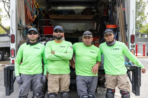 Four landscapers in green shirts standing in front of an open truck filled with gardening tools and equipment.
