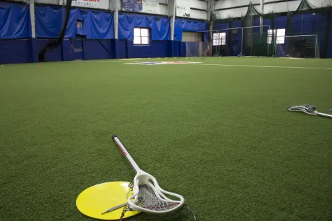 Indoor lacrosse field with green turf, goals, and lacrosse sticks lying on the ground under blue wall padding.