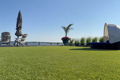 Lawn with patio chairs, folded umbrella, palm plant, flowers, and water view under clear blue sky.