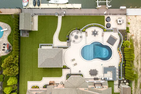 Aerial view of a backyard with a swimming pool, patio seating, dock, boats, and landscaped green lawn by the water.