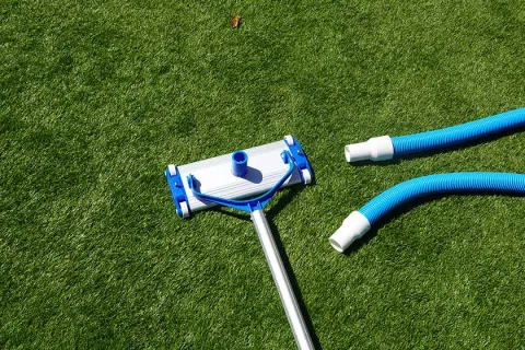 Pool vacuum head and blue flexible hoses lying on green artificial grass outdoor surface.