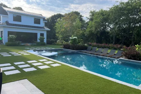 Modern backyard with a swimming pool, lounge chairs, green lawn, and a two-story white house under a partly cloudy sky