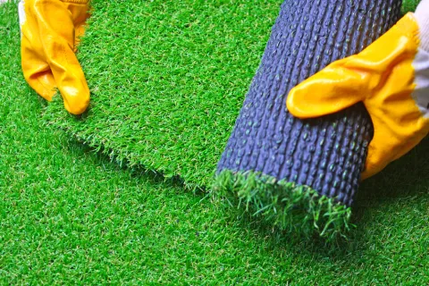 Hands in yellow gloves unrolling green artificial grass turf on a similar turf surface.