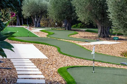 Curved miniature golf course surrounded by olive trees and gravel landscaping on a sunny day