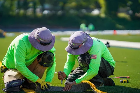 Two workers in green shirts and hats installing artificial turf outdoors on a sunny day.