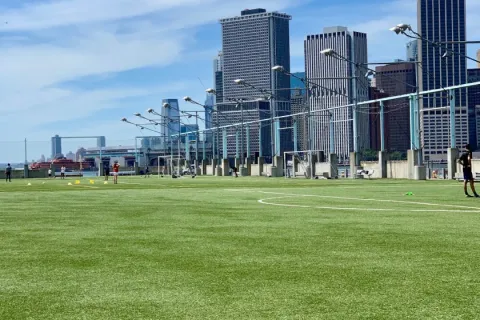 Green soccer field with players practicing, city skyscrapers in background under a blue sky with scattered clouds.