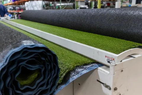 Rolled artificial grass turf being processed in a factory setting with industrial machinery and storage racks.