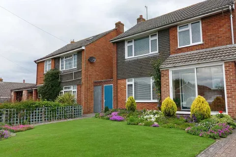 Two attached red brick suburban houses with well-maintained lawns, flowerbeds, and clear windows on a cloudy day