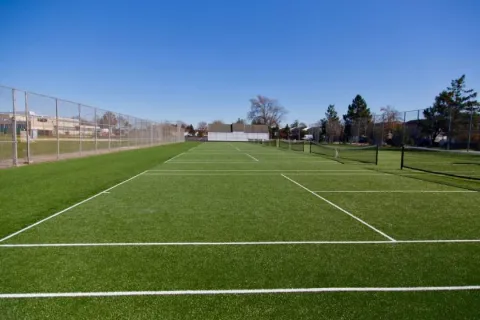 Empty outdoor tennis courts with artificial turf under clear blue sky surrounded by fencing and trees.