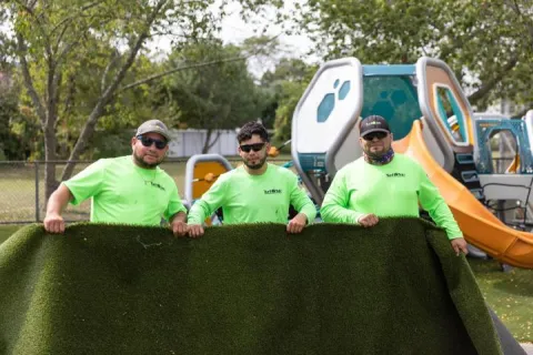 Three workers in neon green shirts holding a rolled artificial grass turf at a playground with slides in background.