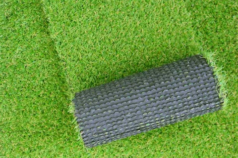 Roll of synthetic artificial grass partially unrolled on green turf surface with visible black backing.