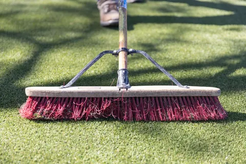 Close-up of a person using a push broom with red bristles to sweep artificial grass outdoors
