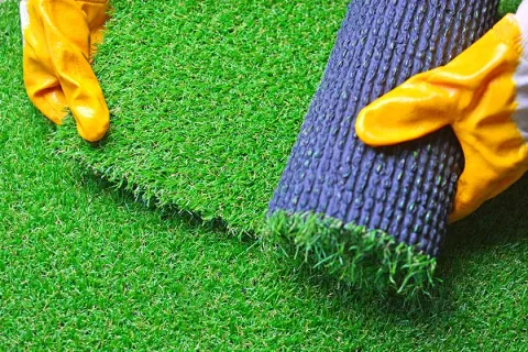Hands wearing yellow gloves laying down and unrolling artificial green grass turf outdoors.