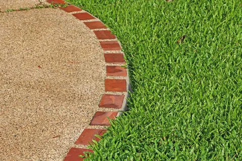 Curved stone walkway edged with red bricks next to vibrant green grass lawn in sunlight.