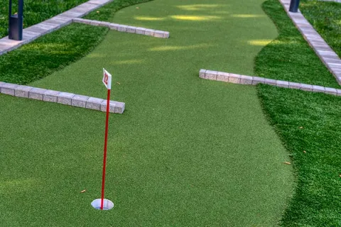 Miniature golf hole 12 with red flagpole and brick course boundaries on green artificial turf.