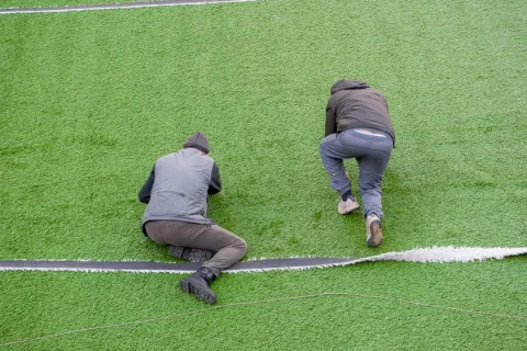 Two workers kneeling on green turf installing or repairing a white line on an artificial grass field.