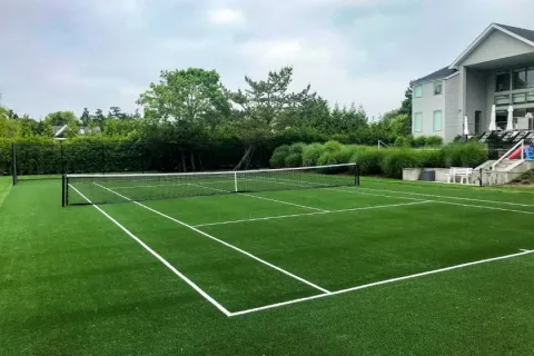 Outdoor tennis court with green artificial turf and white lines next to a modern house and greenery.
