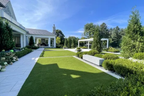 Modern backyard with green lawn, paved walkways, shrubs, trees, and a white gazebo under blue sky.
