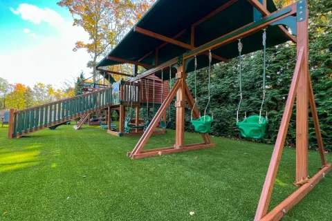 Wooden outdoor playground with green toddler swings, slide, and playhouse under a clear sky and fall trees.