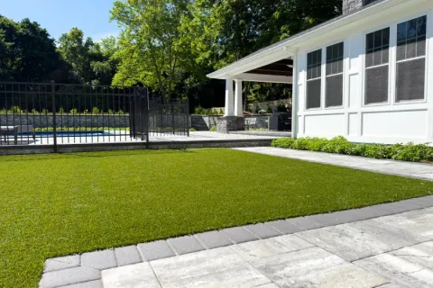 Modern backyard with green artificial turf, stone patio, pool area, and white house with large windows.