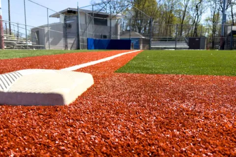 Close-up view of a baseball base on artificial turf with green outfield and sunny sky in background.