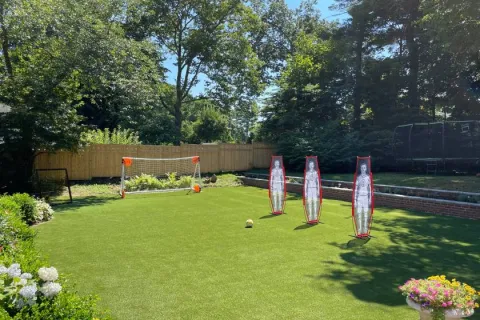 Backyard soccer setup with artificial turf, goalposts, training dummies, and surrounding trees under clear sky.