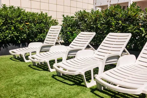Four white plastic lounge chairs on green artificial grass with leafy bushes and a tiled wall in background.