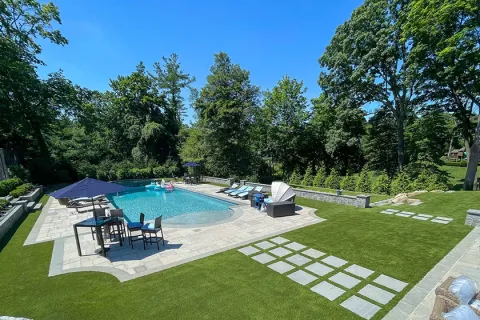 Luxury backyard with swimming pool, lounge chairs, umbrellas, and green lawn surrounded by trees under clear sky.