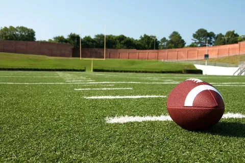 Close-up of a football on the green turf of an empty outdoor football field with goalposts and clear sky.
