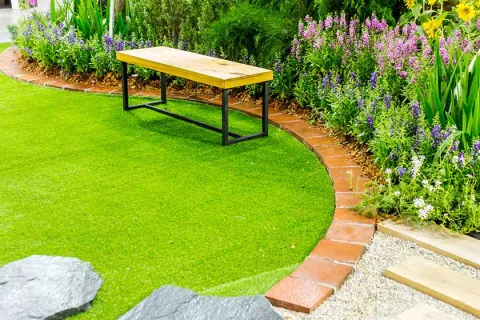 Wooden bench on green lawn with curved brick border and vibrant flower bed in garden landscape