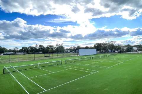 Multiple tennis courts with green artificial turf under a partly cloudy blue sky in a suburban area.