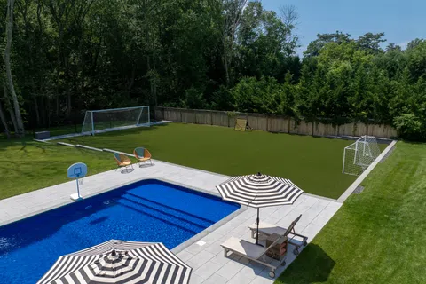 Backyard with blue swimming pool, striped umbrellas, lounge chairs, basketball hoop, and soccer goals on green turf.