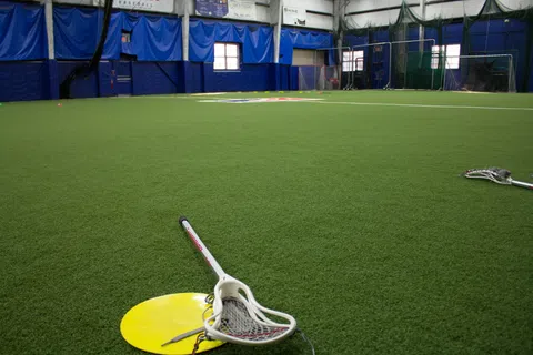 Indoor lacrosse field with artificial turf, lacrosse sticks, and blue padded walls under bright lighting.