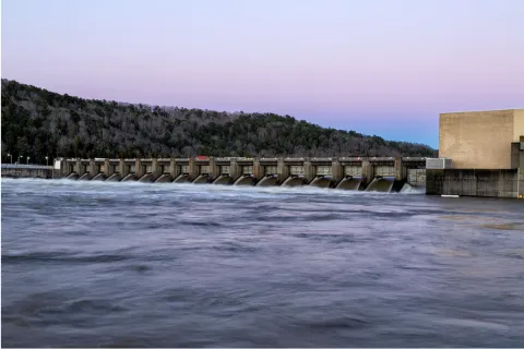 Hydroelectric dam releasing water with forested hill and clear sky at sunset in background.