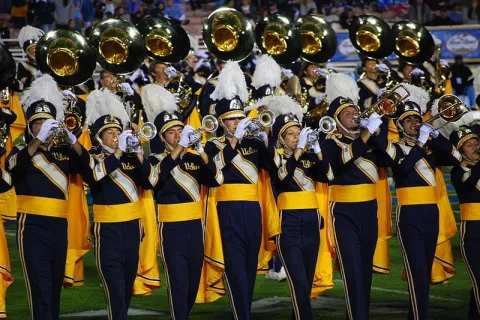 UCLA marching band members performing on field in navy and gold uniforms with brass instruments.