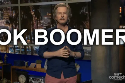 Comedian standing in a studio with the phrase OK BOOMER displayed in large white letters behind him.