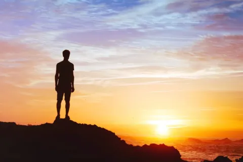 Silhouette of a person standing on rocks watching a colorful sunset over the ocean with clear sky.