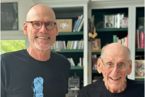 Two men wearing matching black t-shirts with blue footprints, standing inside a room with bookshelves behind them