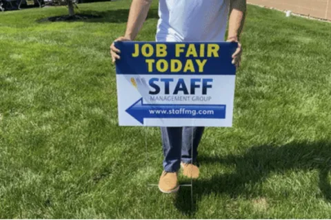 Person holding a job fair today sign on green grass with company name Staff Management Group and website link