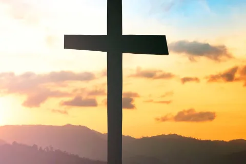 Silhouette of a cross on a hill with a vibrant sunrise and mountain landscape in the background.