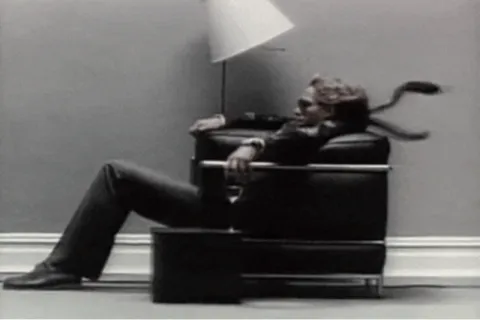 Man in suit and sunglasses reclining on modern black chair against plain wall next to floor lamp