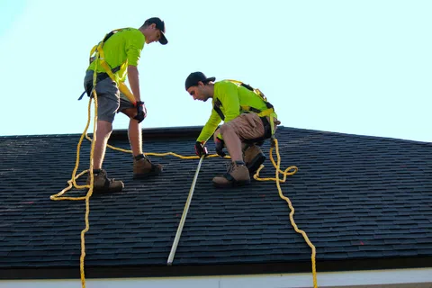 Two roofers in safety harnesses measuring a dark shingle roof on a sunny day with blue sky background