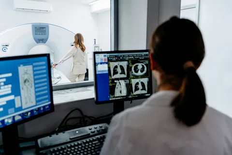 Radiology technician analyzing lung CT scans on computer screens as patient prepares for MRI scan.