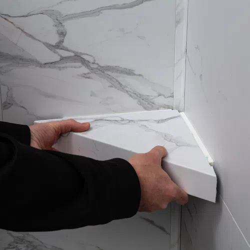 Hands installing a triangular white marble shelf in a corner against matching marble walls.