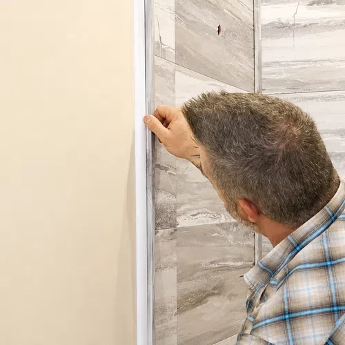 Man installing wallpaper on a tiled bathroom wall near a window wearing a plaid shirt.