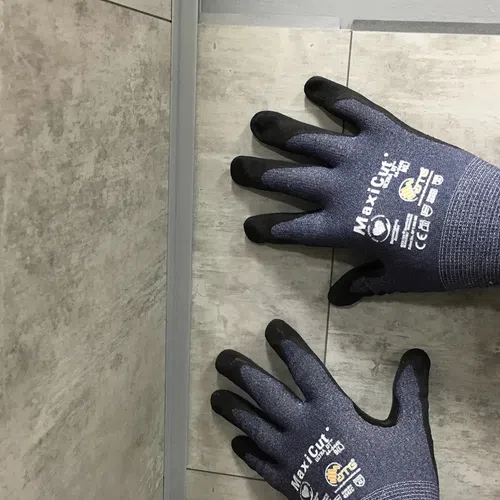 Pair of black and gray MaxiCut safety gloves worn on hands over tiled floor with grout lines.