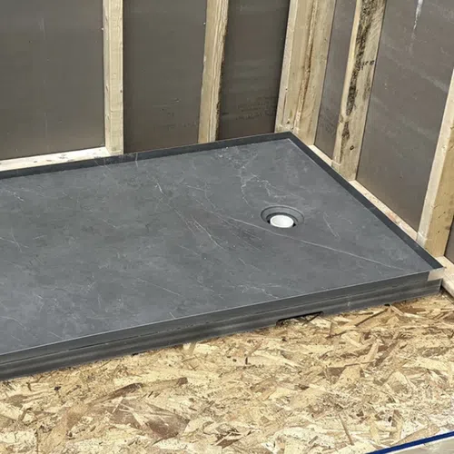 Rectangular dark gray shower base installed on plywood floor in unfinished bathroom with exposed wooden studs.