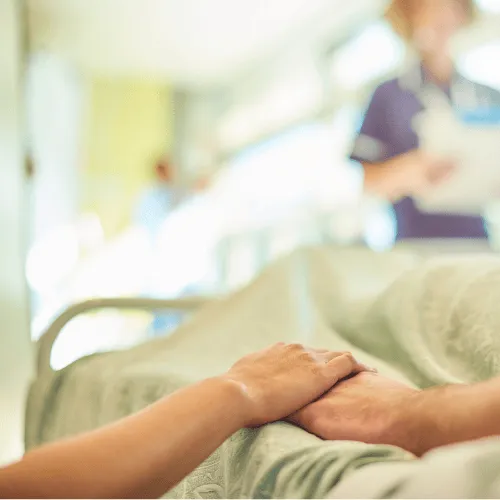 Two hands gently holding above a hospital bed with a nurse in the blurred background.
