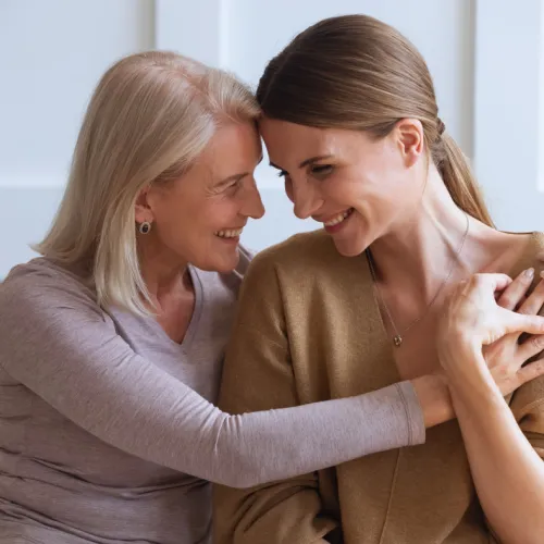 Older woman and younger woman smiling and embracing in a warm, loving moment indoors.