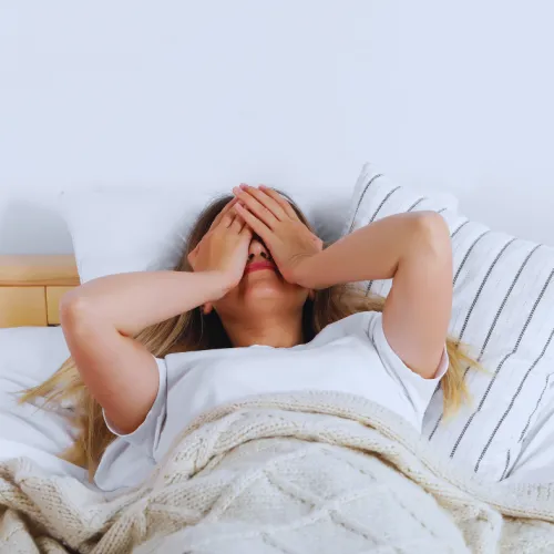 Woman lying in bed with hands covering her face, wrapped in a cozy knit blanket in a bright bedroom.